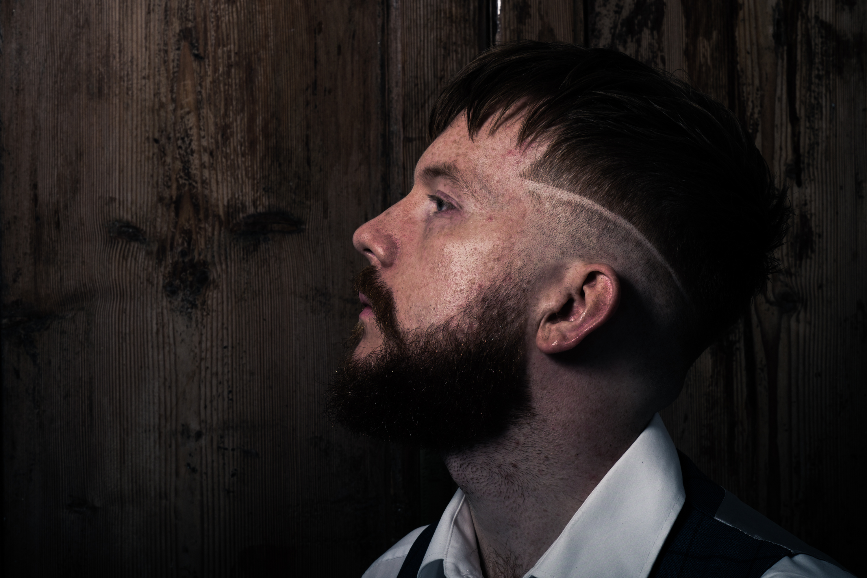A man with a beard wearing a white shirt in front of a wood back drop