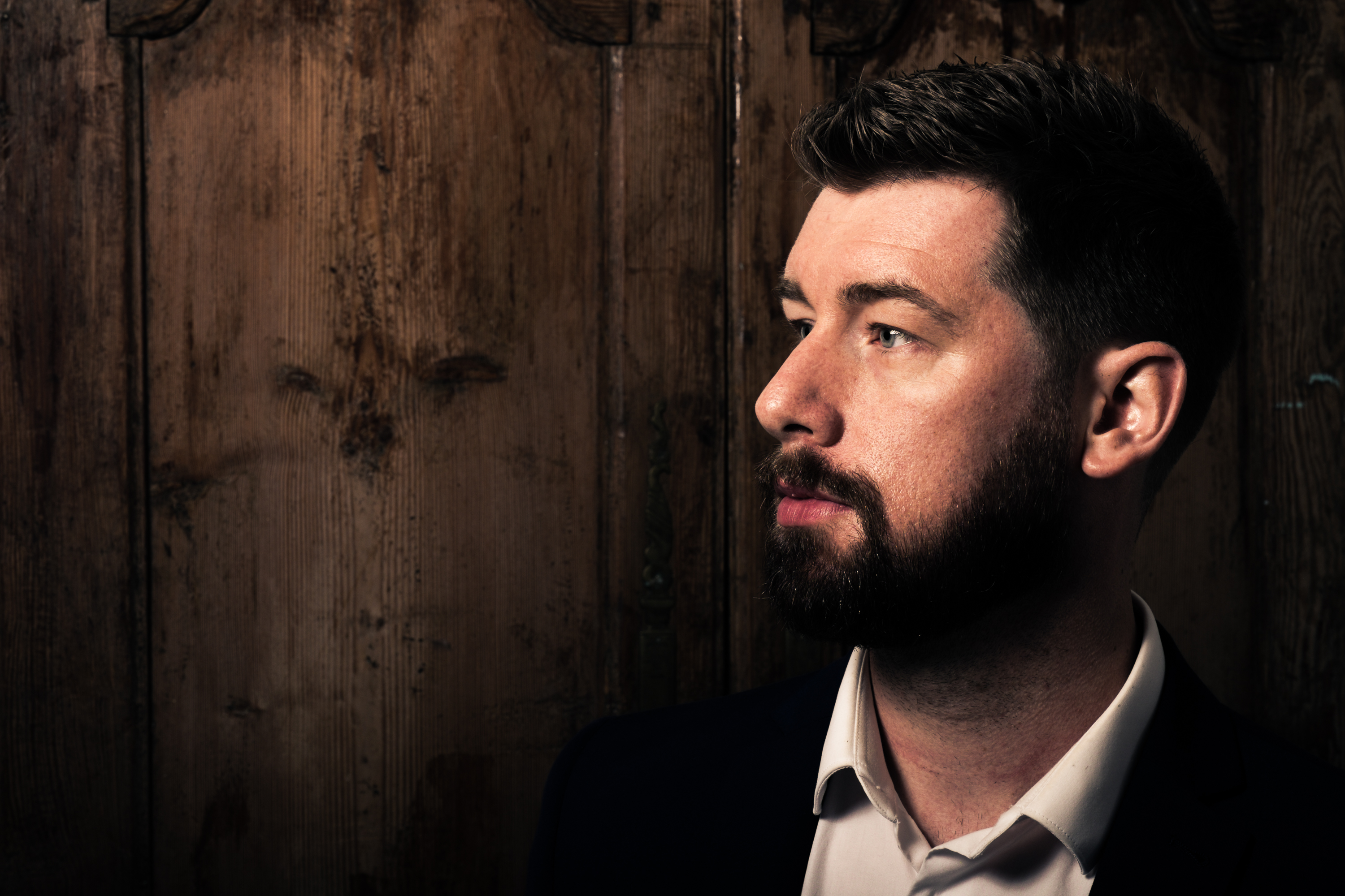 Dark haired man with beard in a suit in front of a wood background