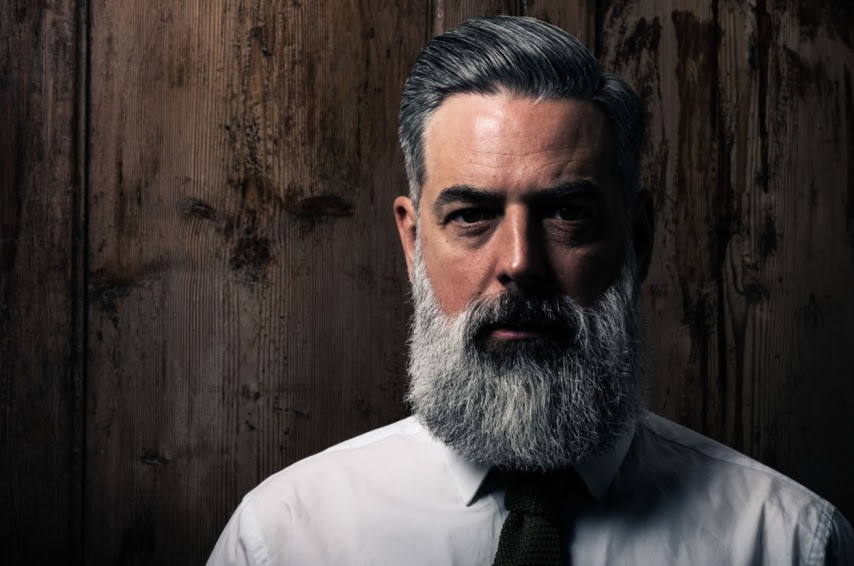 An older man with a grey beard in a suit in front of a wood back ground