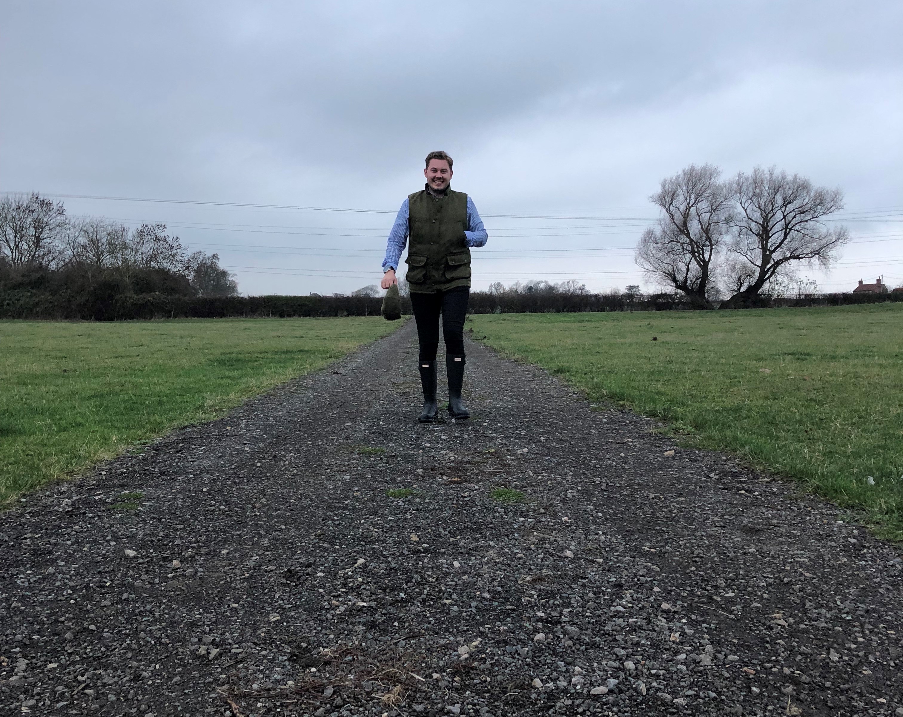 Man wearing gilet and wellies walking through a field on a grey day in the countryside.